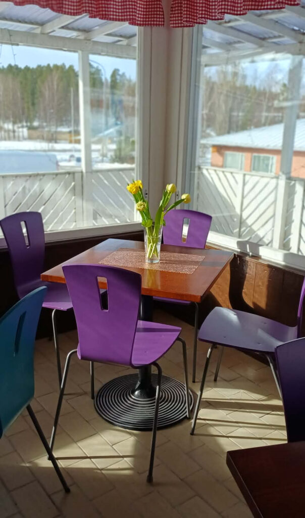 Valoisa ja avara kahvilan sisätila, jossa on värikkäitä tuoleja ja keväisiä kukkia suuren ikkunan äärellä